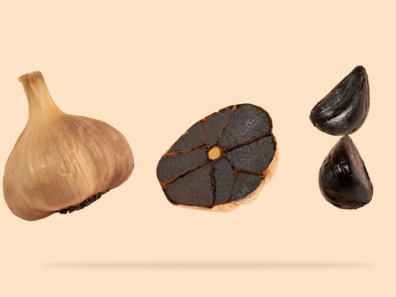 A whole head of black garlic shown next to a sliced head of black garlic and two peeled cloves black garlic, isolated on a light peach background