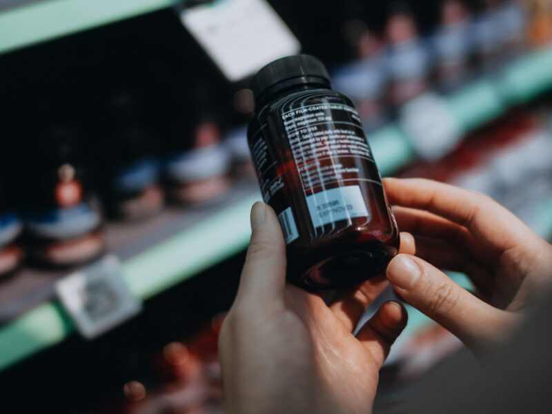 Over-the-shoulder view of hands holding a bottle of probiotic supplements taken off the shelf of a grocery store, reading the label