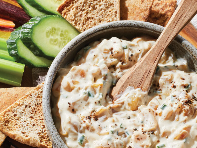 Vegan French Onion Dip in a gray bowl with a wooden spoon on a serving board with pita chips and sliced veggies