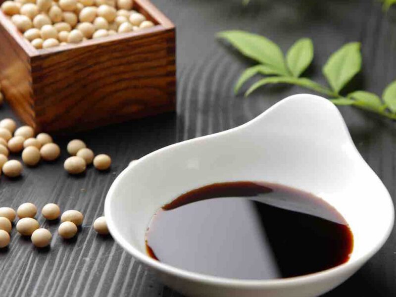 a small box of dried soy beans sits beside a small bowl of soy sauce, with some green leaves on the table behind it