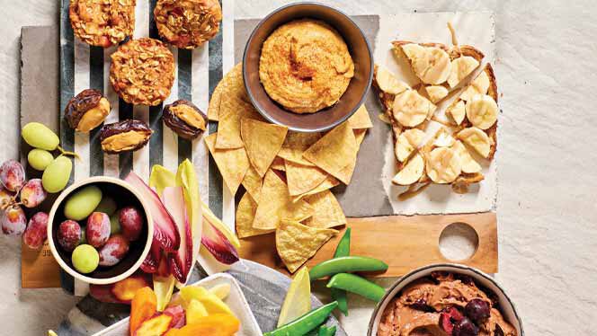 an array of vegan movie night snacks recommended by plant based pros, spread on a striped tablecloth. Snacks include stuffed dates, frozen grapes, snap peas, and banana toast