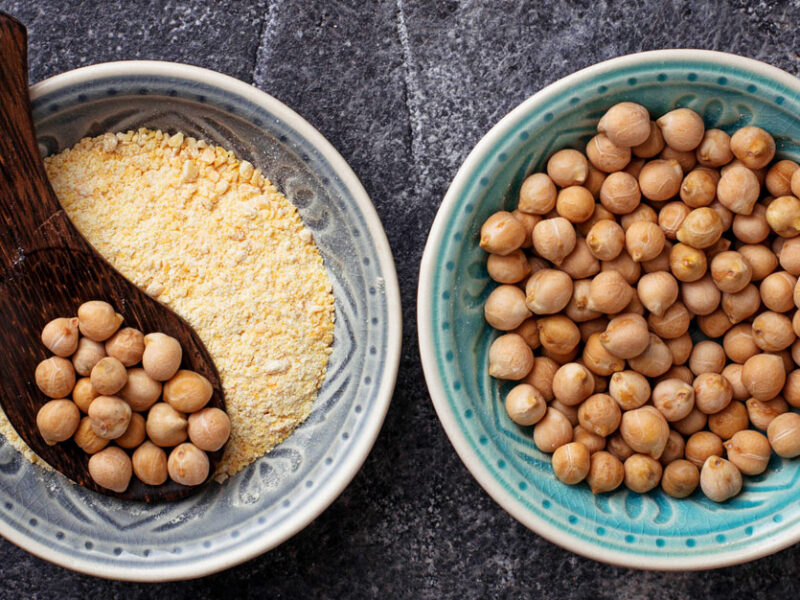 Photo of bowl of ground chickpea flour beside a bowl of whole dried chickpeas, with a spoon full of dried chickpeas resting atop the bowl of chickpea flour