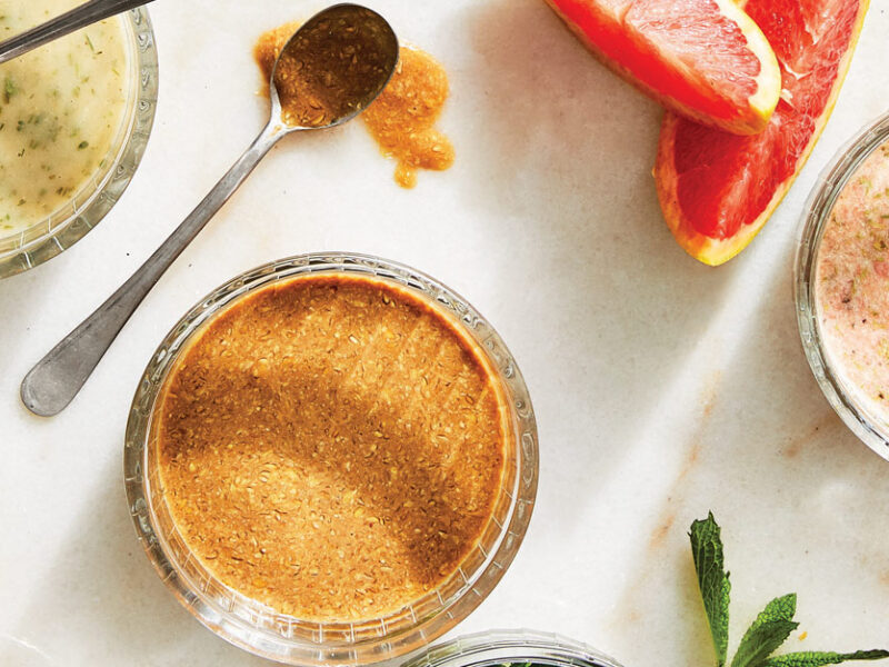 An array of homemade salad dressings made without oil, including vegan ranch, balsamic vinaigrette, and a grapefruit tarragon, in small glass bowls with spoons in them and some of the ingredients shown beside them