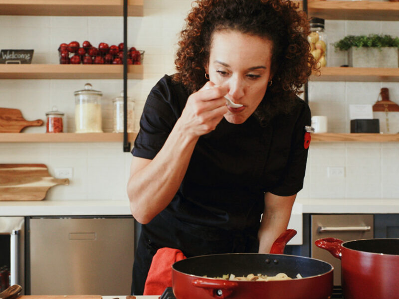 a woman stands over a sauté pan on her stove, with a wooden spoon to her mouth, tasting what she's cooking