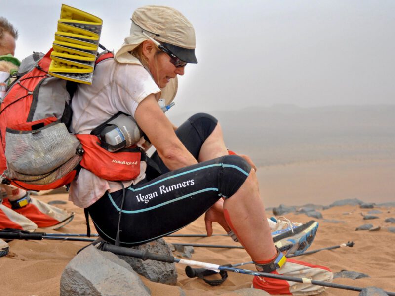 Vegan runner Fiona Oakes adjusts her shoes while wearing a backpack with a fold-up sleeping mat and a scarf around her head as she competes in an ultra marathon. On the side of her leggings is the phrase "Vegan Runners." Photo by Kirsten Kortebein