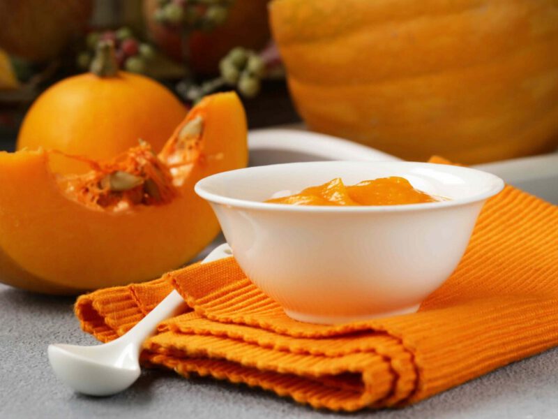 A bowl of pumpkin puree sits atop a folded orange kitchen towel, with cut pumpkin in the background