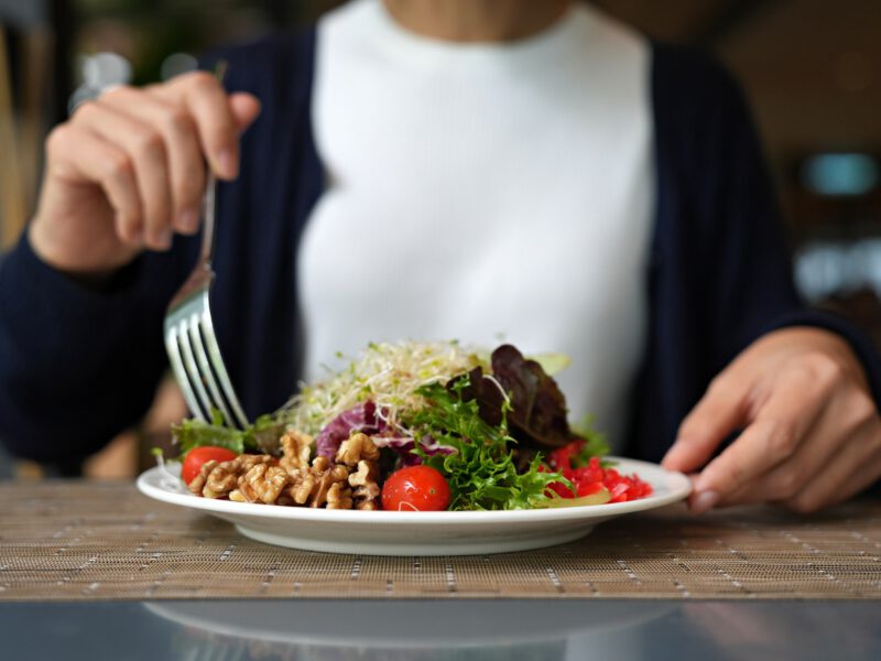 A plate of vegan salad featuring healthy plant-based foods including nuts, greens, and tomatoes, and alfalfa sprouts, with the body of a woman visible sitting behind the plate