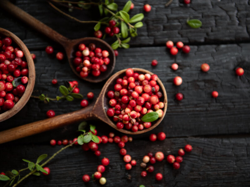 Red cranberries on wooden background, with some in a wooden spoon, and sprigs of their leaves to the side