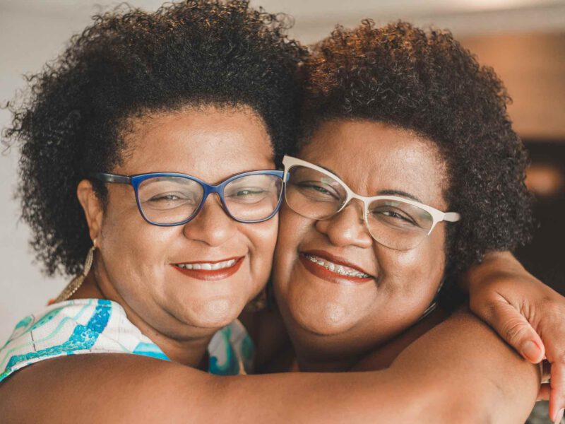 Happy twin sisters who are Black and both wearing glasses hugging, cheek to cheek