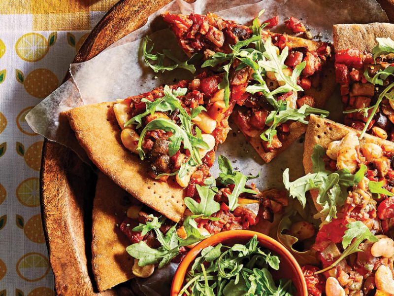 Caponata pizza on a wooden plate on top of a lemon-patterned tea towel