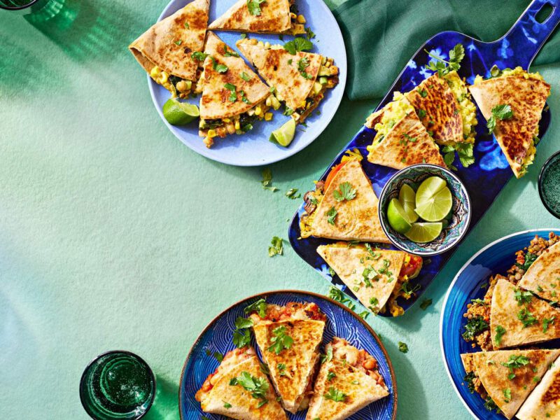 An overhead shot of five different types of vegan quesadillas, on plates on a light blue table