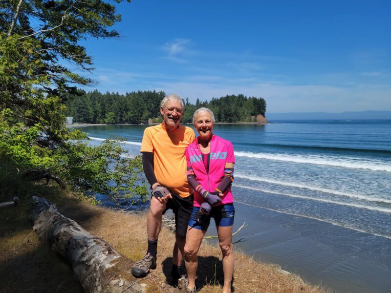 Janet Anspach Rickey and her husband stand on the shore of a lake, wearing cycling outfits, looking healthy after adopting a WFPB sos-free diet for weight loss and heart health
