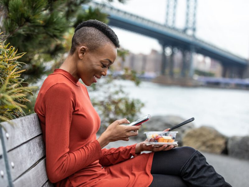 A young Black woman sits by the river in New York City eating healthy plant-based food