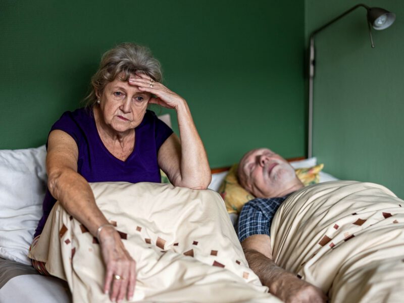 An older white woman sits up in bed, unable to sleep, while her husband lies snoring next to her