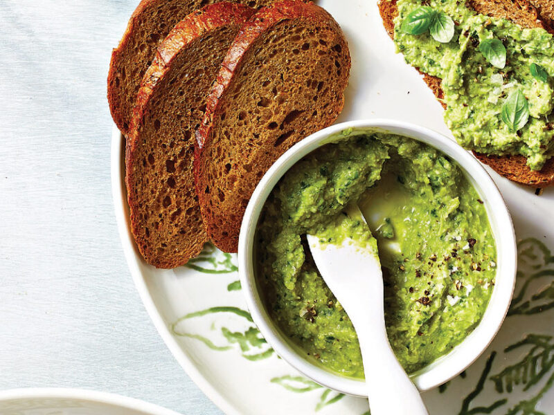 Pesto Artichoke Spread in a small white bowl with a serving knife on a plate with slices of whole wheat bread