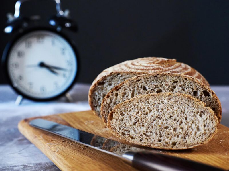A loaf of sliced whole wheat bread sits on a wooden cutting board with an old alarm clock in the background