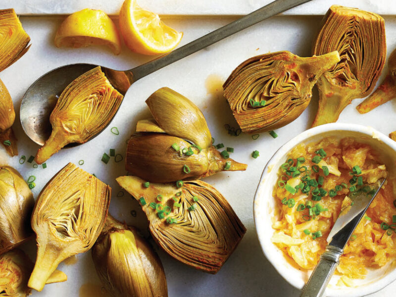 Lemon-Garlic Roasted Artichoke halves on a white platter next to a white ramekin of pressed garlic