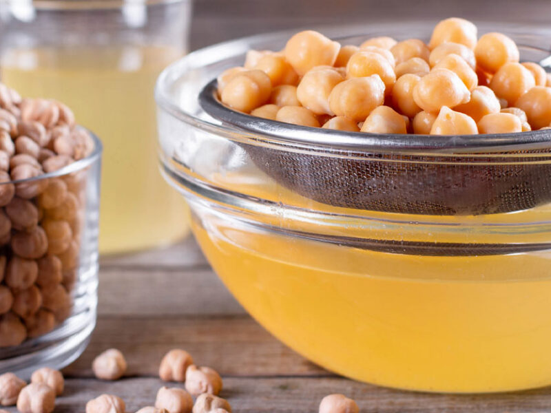 A mesh strainer full of chickpeas is placed over a glass bowl where aquafaba has drained down into it. A glass of dried chickpeas sits to the lefthand side.