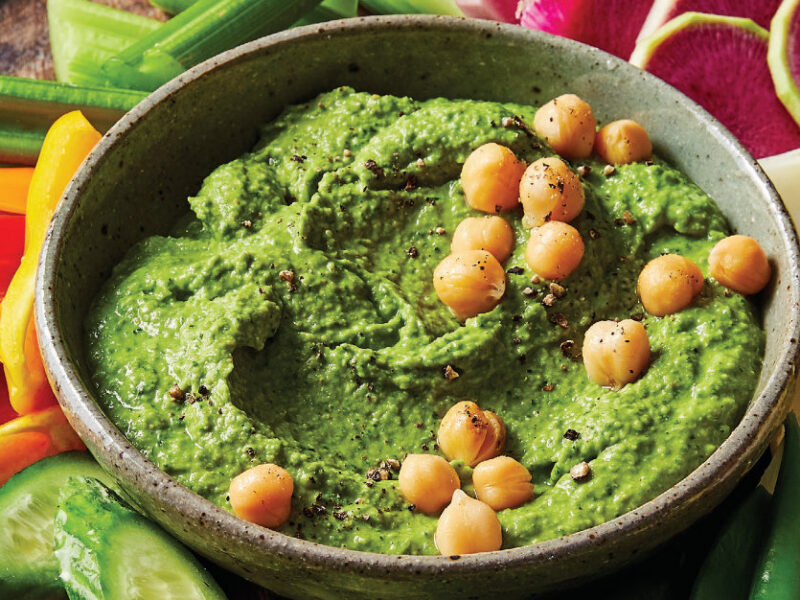 Fresh Spinach Hummus garnished with whole chickpeas on a round platter surrounded by bite sized cucumber, celery, radish, and bell pepper