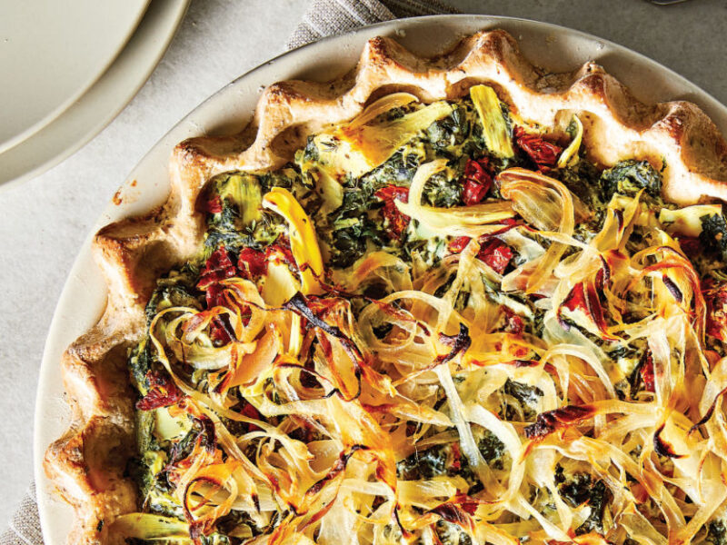 Quiche Florentine with Artichoke Hearts and Sun-Dried Tomatoes on a white plate on a white tablecloth with a beige and white striped napkin and a couple of empty plates
