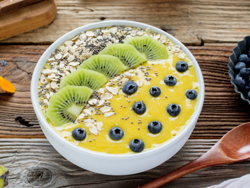 Mango Smoothie Bowl on a rustic wooden table next to a bowl of fresh blueberries, fresh turmeric root a banana and a wooden spoon