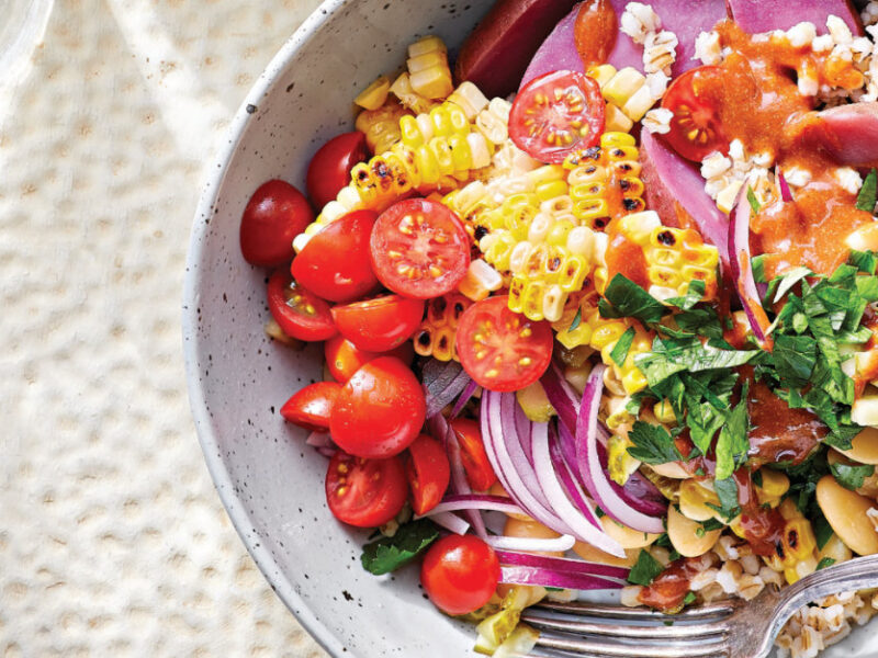 Rainbow Veggie Barley Bowl in a white pottery bowl on a white background next to a small glass of lemon slices