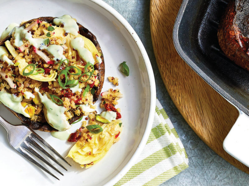 Quinoa- and Artichoke-Stuffed Mushrooms on a white plate sitting on a green and white striped cloth napkin next to a skilleet with one grilled portobello mushroom sitting face up, ready to be stuffed