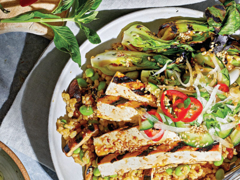 Grilled Bok Choy, Tofu, and Mushrooms with Brown Rice and Ponzu Sauce on a white plate with a sprig of Thai basil on a wooden chopping board