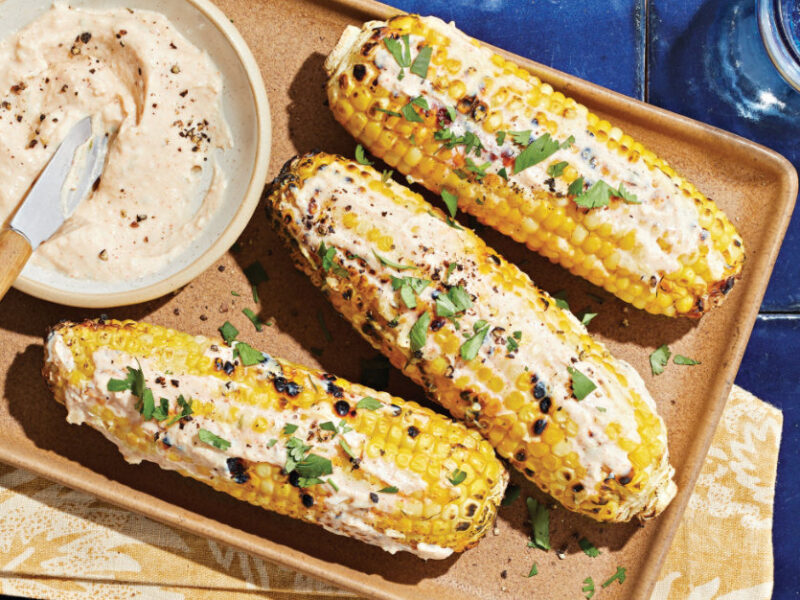 Three cobs of Vegan Elote (Mexican Street Corn) on a platter with a small bowl of lime-infused crema