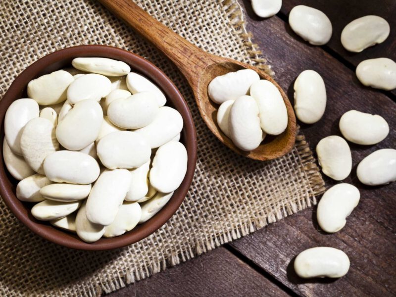 Top view of a brown bowl filled with lima beans shot on rustic wood table. A wooden spoon is beside the bowl with some beans on it.