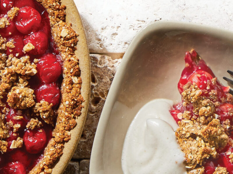 Cherry Crumble Pie in a round brown pie dish next to a plate with a serving of the pie and a dollop of Aquafaba Whipped Cream