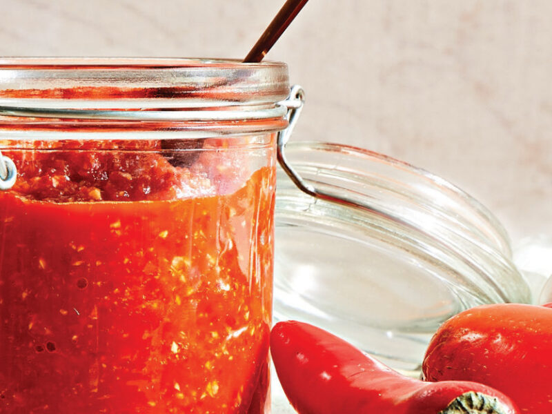 A mason jar of Chile-Garlic Sauce with a teaspoon resting inside, next to red chile peppers and raw garlic
