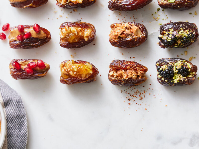 An array of stuffed dates, stuffed with five different fillings, with a small bowl of nut butter and pomegranate arils shown off to the side