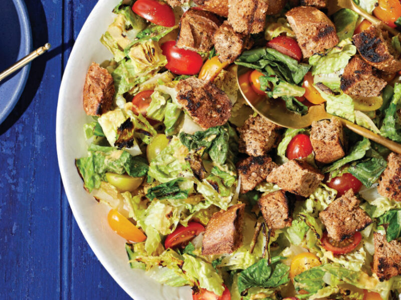 Grilled Caesar Salad in a white bowl with woode serving spoons on a bright blue table next to a small blue ceramic bowl of dressing