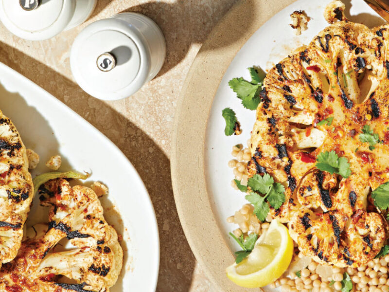 Harissa Cauliflower Steaks with Date Couscous on a white plate next to a salt and pepper shaker and a plate of cauliflower steaks