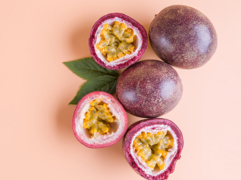 Two whole passion fruit beside three cut-open halves of passion fruit showing the colorful pulp inside, on top of a plain light pink background