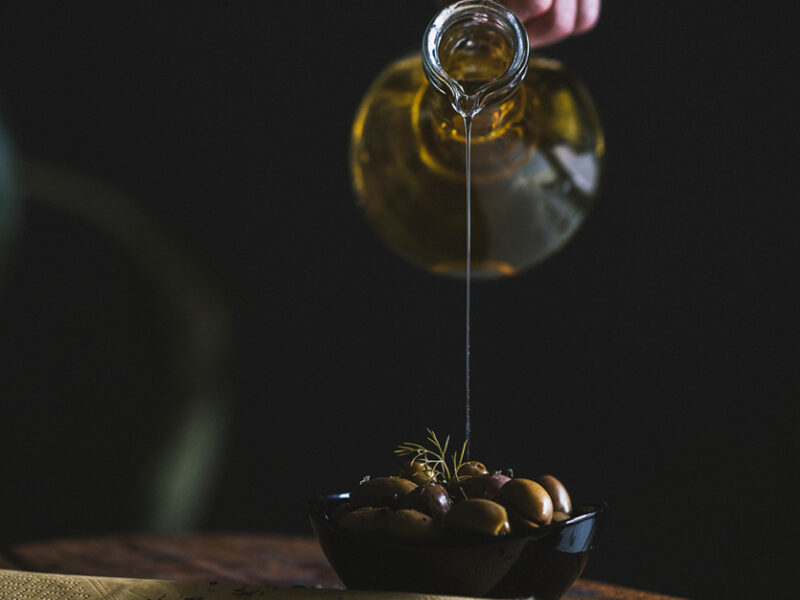 olive oil being poured from a carafe into a small bowl of olives, with a dark background
