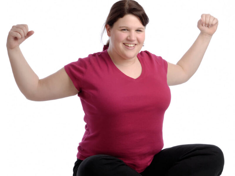 happy overweight young woman sits crosslegged on the floor with her arms lifted out to the side
