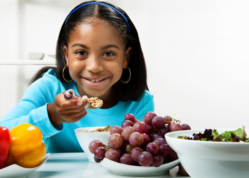 young girl sitting at a table eating healthy food, in front of a bowl of grapes and a bowl of greens