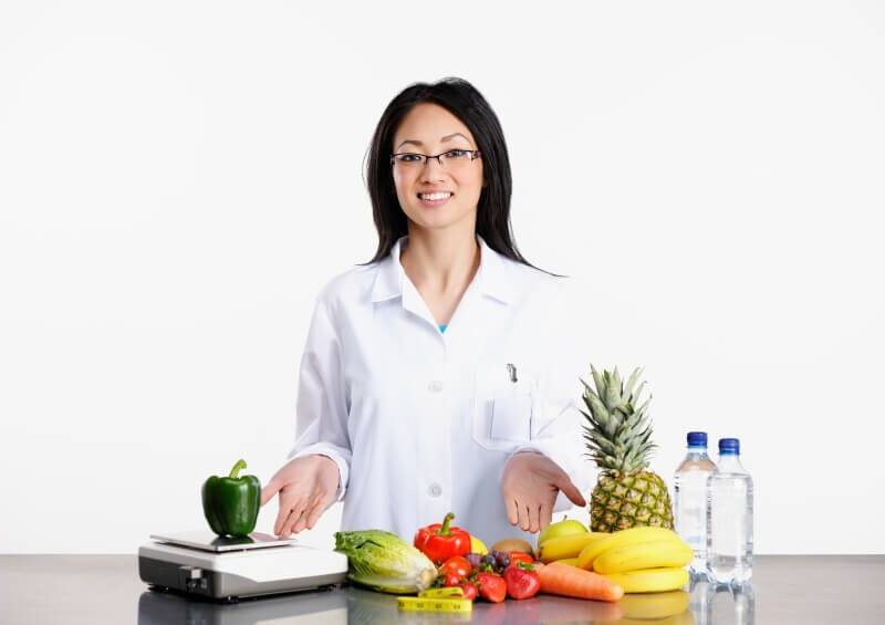 A female doctor in a white lab coat with her hands outstretched in front of her towards a colorful array of fresh fruits and veggies