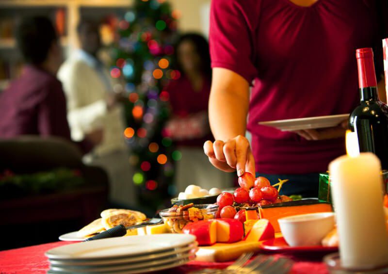 A hand reaches out to grab a grape from a holiday table which also has a block of cheese, a candle, and a bottle of wine