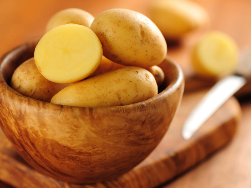 A wooden bowl of raw potatoes with a knife in the background