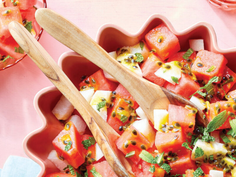 Watermelon Salad with Passion Fruit Dressing in a pink bowl on a pink background with wooden serving spoons in the bowl
