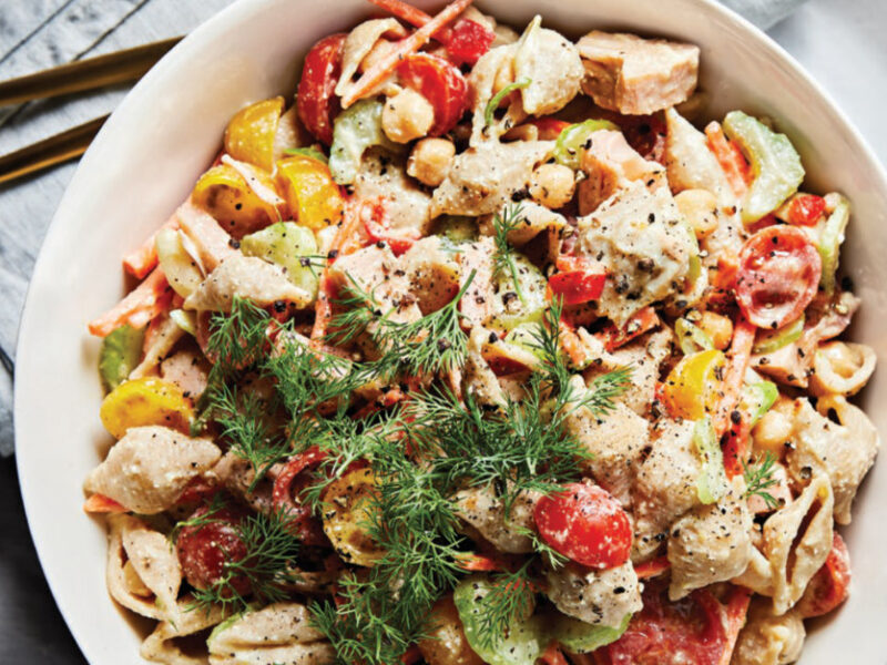 Shellroni Salad with Jackfruit, Cherry Tomatoes, and Creamy Cashew Dressing in a white bowl sitting on a white plate on a white tablecloth, with a garnish of fresh dill on top