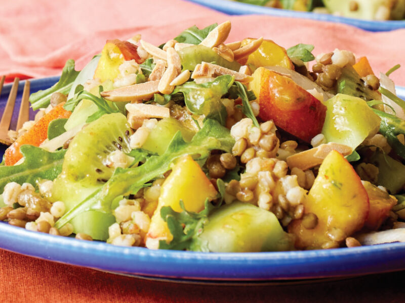 A serving of Honeydew, Peach, and Barley Salad on a blue dinner plate on an orange table top with a pink cloth napkin