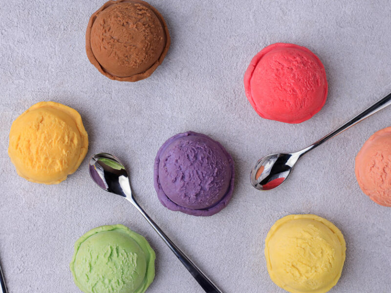 Colorful individual scoops of ice cream on a light purple surface, with spoons next to them