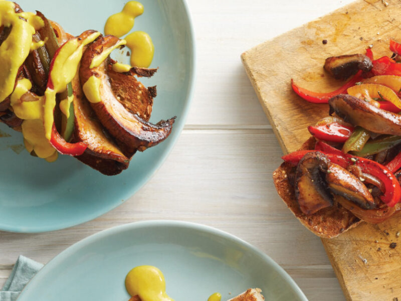 Mushroom Cheesesteak Sandwich on a ceramic plate next to a chopping board with two more sandwiches on it on a white wooden table