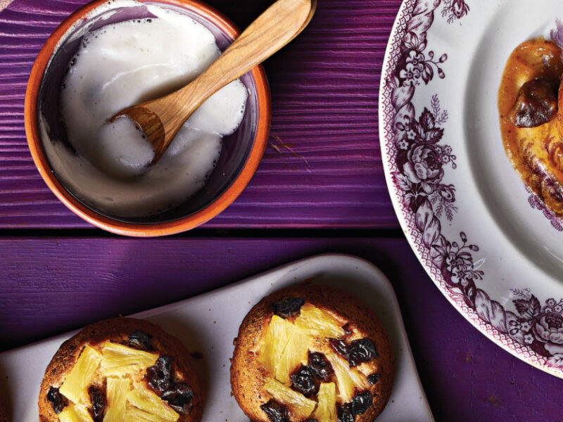 A Upside-Down Pineapple Cupcake on a white and purple tea plate on a purple wooden table next to a small bowl of aquafaba and the corner of a muffin tray showing three cupcakes