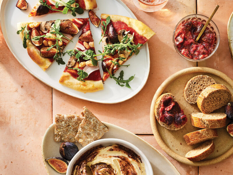 A table spread with a variety of fresh fig dishes: a pizza topped with fresh figs, hummus in a ramekin with a fig paste swirled in, fig jam spread on whole grain bread, a fig and apple salad, and maple-roasted figs