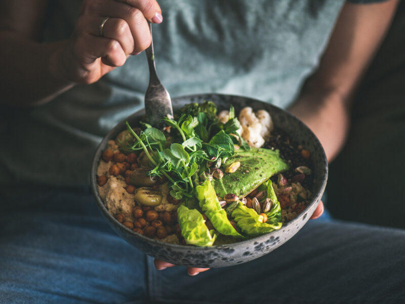 person eating a healthy vegan bowl meal featuring hummus, vegetable, salad, beans, couscous and avocado